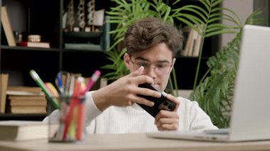 Odaklanmış erkek öğrenci cep telefonu oyunu oynuyor masada dizüstü bilgisayar ve kırtasiyeyle oturuyor. Adam evde sınava hazırlandıktan sonra ürperiyor.