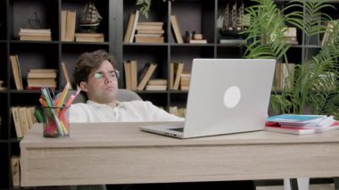 Adam koltuğa uzanıp dizüstü bilgisayarını rahatça izliyor. Kompozisyon masa ögeleri ve arkaplan dolusu kitap ve yeşillik içerir