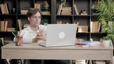 Üniversite öğrencisi rahat kütüphanede dizüstü bilgisayarda özenle çalışır. İnsan, kitaplar ve doğal ışık ile çevrili olarak oturur ve verimli bir atmosfer yaratır.