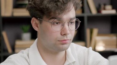 Gözlüklü genç adam kütüphanede okuyor ve akademik uğraşlara odaklanıyor. Öğrenci cevap ararken gülümsüyor.