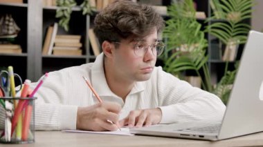 Genç adam dizüstü bilgisayara yazıp çevrimiçi eğitim sırasında bilgisayar ekranına bakıyor. Ev eğitimi alanında kitaplar ve ahşap mobilyalar vardır.