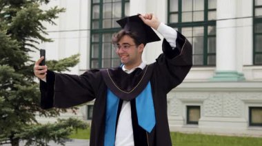 Heyecanlı mezun adam havan topunu eğip telefon açısını değiştiriyor. Önlüklü öğrenci, ladin ağacıyla klasik binanın yakınında poz veriyor