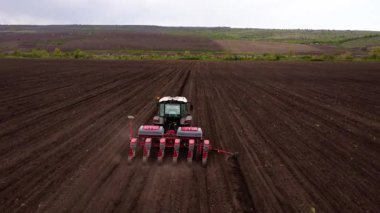 Tarım makinesi geniş kahverengi alanda çalışır ve toprakta paralel çizgiler oluşturur. Çekici, yerleştirme sürecini genişletmek için hareket eder