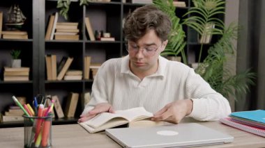 Odaklanmış üniversite öğrencisi final sınavlarına hazırlanırken ders kitabı okur. Adam, hafif ev kütüphanesinde dizüstü bilgisayar ve defterlerle masada oturuyor.