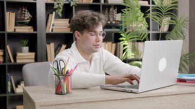 Üniversite öğrencisi rahat ev ofisinde uzaktan eğitim için dizüstü bilgisayar kullanarak masada oturuyor. Kitaplık eğitim atmosferine sıcaklık katıyor