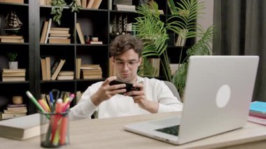 Genç adam cep telefonuyla bilgisayar ve kitaplarla ofis masasında oyun oynuyor. Sosyal medyada vakit harcamayı erteliyor