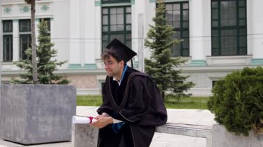 Önlüklü genç erkek mezun üniversitenin önündeki bankta oturuyor. Adam, gelecekteki işiyle ilgili sevinç ve heyecanla gülümseyen bir diplomaya sahip.