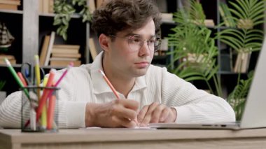 Brunet 'li adam elinde kalemle dizüstü bilgisayara odaklanmış bir şekilde bakıyor. Ofiste yeşil bitkiler ve renkli kalem tutacağı yakınındaki kitaplar yer alıyor.