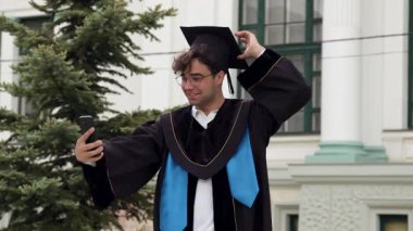 Mezun adam akademik cübbeyle selfie çekerken gülümsüyor. Öğrenci, yüksek binanın önünde duran havan tahtasına hafifçe uyum sağlıyor.