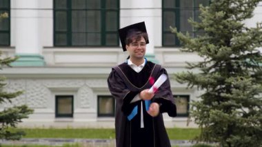 Gecelik ve kepli mutlu üniversite mezunu mezuniyeti üniversitenin önünde diplomasını tutarak kutluyor. İnsan gururla ve kendinden emin durur.