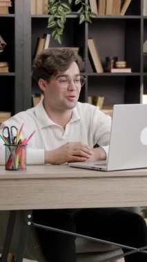 Erkek öğrenci çevrimiçi oturum sırasında masada el kol hareketi yaparken konuşuyor. Yeşil bitkiler ve uzun kitaplık görsel çerçeve olarak arkasında durur