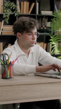 Genç adam dizüstü bilgisayarda yazar ve çalışırken ekrana odaklanır. Kitapları ve bitkileri olan modern kitaplık ahşap masanın arkasında duruyor.