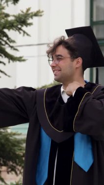 Genç mezun, üniversite binasının önünde akademik elbise ve havan topu ile selfie çekiyor. Adam mezuniyeti kutluyor.