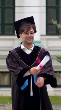 Gecelik ve kepli mutlu üniversite mezunu mezuniyeti üniversitenin önünde diplomasını tutarak kutluyor. İnsan gururla ve kendinden emin durur.