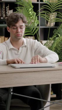 Genç adam organize çalışma alanındaki ahşap masada otururken dizüstü bilgisayarı açıyor. Kitaplık dolusu kitap yavaş çekimde bilgi arzusunu gösteriyor