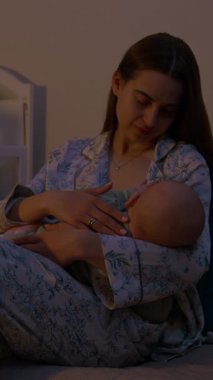 Rahat pijama giyen genç anne yeni doğmuş bebeği koltukta oturarak emziriyor. Anne ve bebek huzurlu bir ortamda loş bir odada.