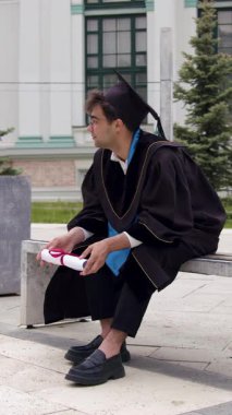 Şapka ve cübbe giyen düşünceli mezun üniversitenin dışında bankta oturuyor diplomasını tutuyor ve mezuniyetten sonra kariyer yapmayı düşünüyor.