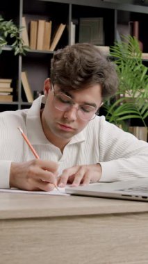 Genç erkek öğrenci çevrimiçi öğrenmek için dizüstü bilgisayar kullanarak not alır. Adam kitaplık ve bitkilere karşı resepsiyondan çalışıyor.