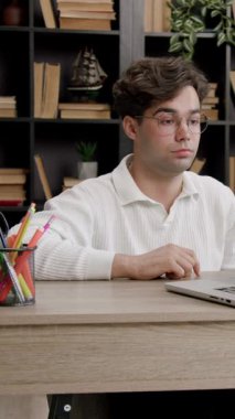 Brunet 'li adam dik oturur ve çevrimiçi çalışırken dizüstü bilgisayara dikkatle bakar. Parlak ışık, kitaplara ve bitkilere karşı işyerlerini aydınlatıyor