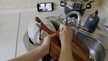 Ev hanımı kesme tahtasını yıkıyor mutfaktaki akıllı telefondan dadı kamerası çevirisini izliyor. Anne evde bulaşık yıkamayı kontrol ediyor.