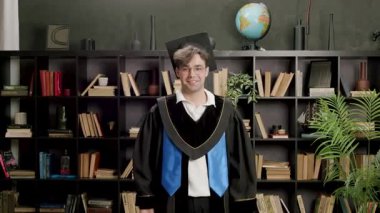 Mezuniyet elbisesi ve havan tahtasındaki genç adam diplomasını gururla taşıyor. Öğrenci başarıyı kollarını kaldırıp kütüphanede gülümseyerek kutluyor.