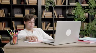 Genç bir öğrenci sandalyeye yaslanıyor ve ciddi bir suratla dizüstü bilgisayara bakıyor. Modern odada kitaplar ve bitkiler varken kalem elinde duruyor.