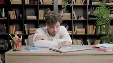 Adam kitap ve klasörlerle dolu masada otururken gözlere ve yüzlere el masajı yapıyor. Duruş zihinsel yorgunluk ve yansıma anını gösterir.