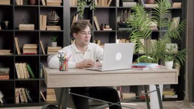 Erkek öğrenci çevrimiçi oturum sırasında masada el kol hareketi yaparken konuşuyor. Yeşil bitkiler ve uzun kitaplık görsel çerçeve olarak arkasında durur