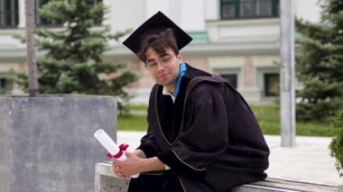 Yüzünde hüzün belirtileriyle mezun olmuş, bankta oturmuş gülümsüyor. Adam diplomasını sarmış ve üniversite kampüs parkına bakıyor.