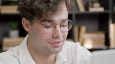 Gözlüklü akıllı adam gülümseyerek çalışırken dizüstü bilgisayara bakıyor. Geçmişi kitaplarla bulanık ve insan yüzünün arkasındaki yumuşak ışık