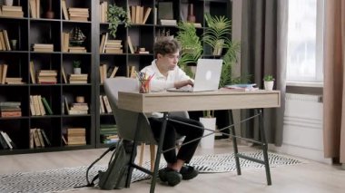 Odaklanmış öğrenci, arka planda sırt çantası ve kitaplıklarla ev ofisinden ders çalışırken dizüstü bilgisayar kullanıyor. Adam doğal pencere ışığından hoşlanıyor.