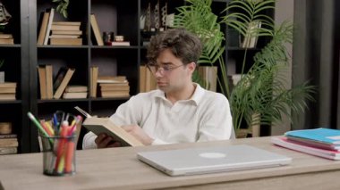 Yoğunlaşmış erkek öğrenci, gözlük takarak kalın ders kitapları okur. Adam, kitaplıklarla çevrili dizüstü bilgisayar ve kırtasiyeyle masa başında çalışıyor.