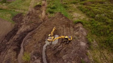 Kazıcılar toprak taşıma operasyonları sırasında inşaat alanında yeşillik olan çamurlu bölgede çalışırlar. Makineler siper şekillendirme manzarası kazıyor