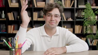 Kendine güvenen erkek öğrenci kütüphanenin merkez ofisinde masa başında otururken elini kaldırıyor. Soruya cevap vermeye çalışan zeki ve dost canlısı birine benziyor.