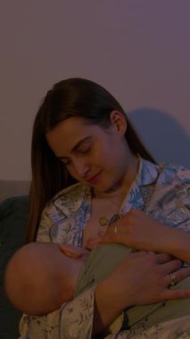 Anne sevgiyle yeni doğmuş bebeği emziriyor koltukta rahat bir şekilde oturuyor. Genç aile, evde barışçıl ve samimi bir bağ kurma anını resmediyor.