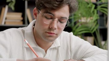 Gözlüklü adam derslerine odaklanıyor kalemle notlar alıyor. Öğrenci kitaplarla dolu kütüphanede çalışıyor. Huzurlu bir alan yaratıyor.