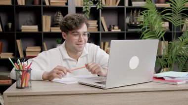 Adam internette konuştuktan sonra gülümser ve sandalyeye yaslanır. Masada kitapların düzenli arkaplanlı dizüstü bilgisayar ve kırtasiye malzemeleri var