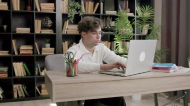 Genç adam dizüstü bilgisayarda yazar ve çalışırken ekrana odaklanır. Kitapları ve bitkileri olan modern kitaplık ahşap masanın arkasında duruyor.