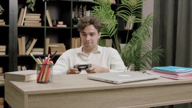 Masada bilgisayar oyunu oynayan genç bir adam var. Öğrenci gülümsüyor. Çalışma odasında oturuyor. Etrafı kitaplar ve bitkilerle çevrili.