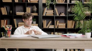 Masadaki ciddi öğrenci kalemi dikkatle inceliyor. Adam sakin kalıyor ve bilgisayarla masa başında kısa bir ara vermeye odaklanıyor.