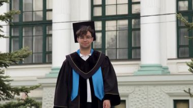 Mezun olan adam klasik binanın yanında mutlu bir yüzle duruyor. Genç öğrenci güvenle el ele tutuşuyor, akademik bir elbise giyiyor ve gülümsüyor.