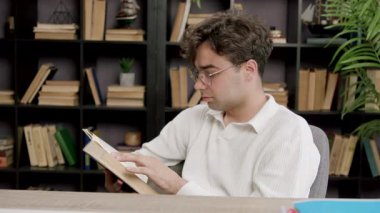 Genç üniversite öğrencisi hafif ev kütüphanesinde ders kitabı okuyor. İnsan, kitaplarla çevrili rahat ve sessiz bir ortamda çalışmalara odaklanır.