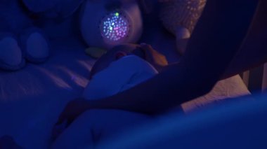 Annenin elleri, gecenin bir yarısı beşiğinde uyanan küçük bebeğe sarılır. Bakıcı kadın, evdeki yarı karanlık yatak odasında küçük çocuğu rahatlatıyor.