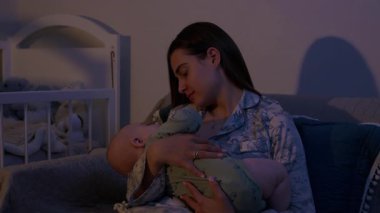 Gevşek saçlı genç bir kadın koltukta oturan sevimli bebeği nazikçe kucaklıyor. Anne, çocuk gece geç saatte uyandığında onu teselli eder.