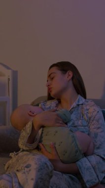 Genç bir kadın, klasik beşiğin yanındaki rahat koltukta oturan şirin bir bebeği beşikte tutuyor. Sevgi dolu anne sakin ol bebek çocuk gece yatak odasında ninni söylüyor.