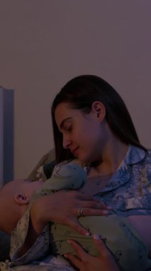 Gevşek saçlı genç bir kadın koltukta oturan sevimli bebeği nazikçe kucaklıyor. Anne, çocuk gece geç saatte uyandığında onu teselli eder.