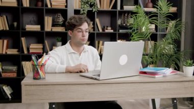 Brunet 'li adam dik oturur ve çevrimiçi çalışırken dizüstü bilgisayara dikkatle bakar. Parlak ışık, kitaplara ve bitkilere karşı işyerlerini aydınlatıyor