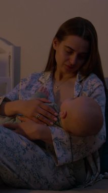 Genç anne kanepede oturan yeni doğmuş bebeği şefkatle emziriyor. Pozitif kadın, loş ışıklandırılmış yatak odasında bebekle yakınlaşmaktan hoşlanır.