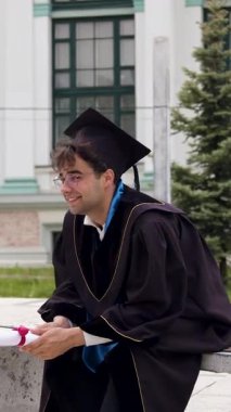 Önlüklü genç erkek mezun üniversitenin önündeki bankta oturuyor. Adam, gelecekteki işiyle ilgili sevinç ve heyecanla gülümseyen bir diplomaya sahip.