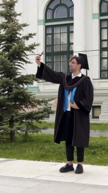 Mezuniyet elbisesi ve havan topu giymiş gururlu bir öğrenci üniversite binasının önünde akıllı telefonuyla selfie çekiyor. Adam mutlu görünüyor.